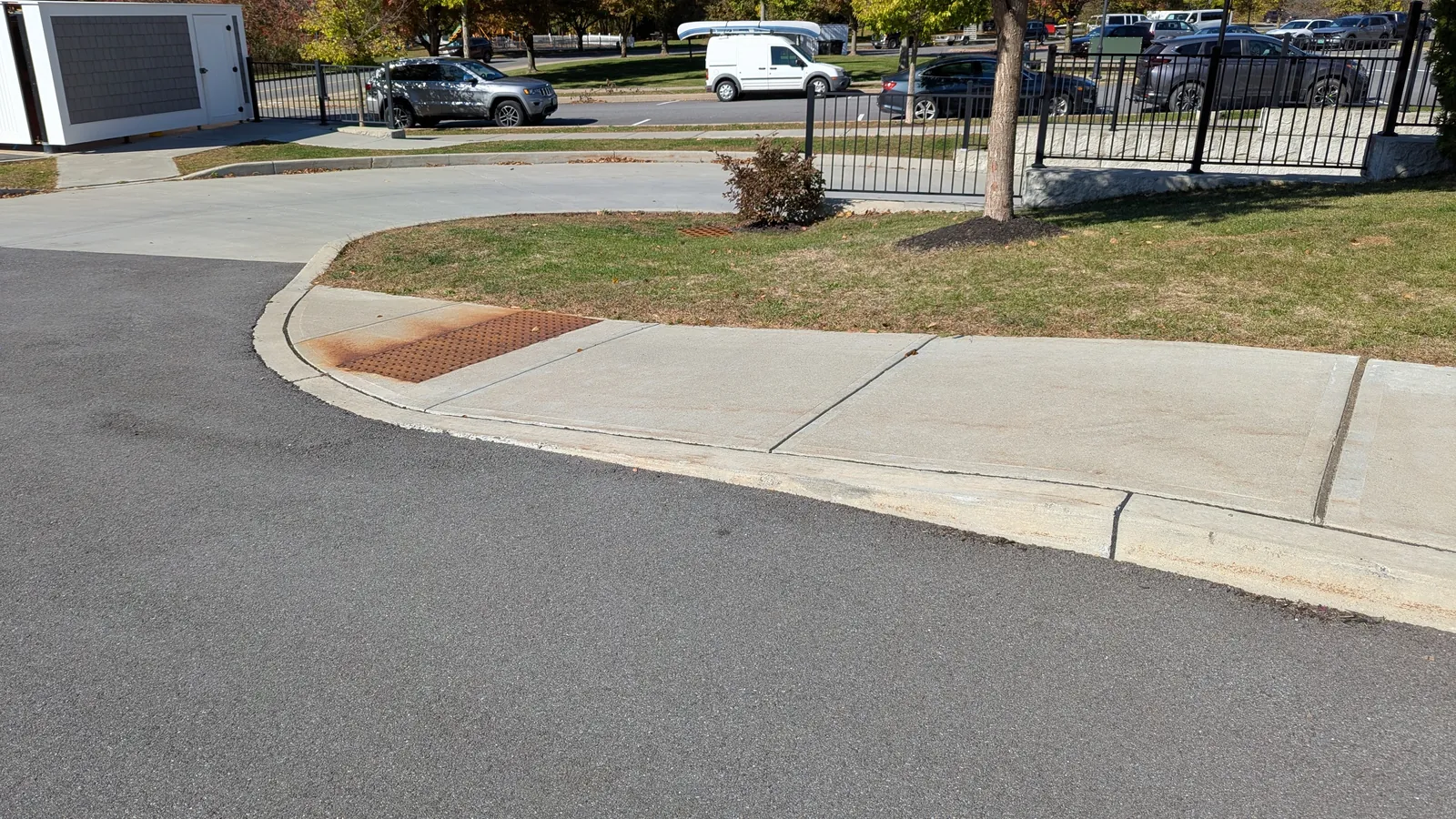 Concrete curb ramp with a red-brown warning panel at a lowered curb beside an asphalt drive, a fenced parking lot behind it
