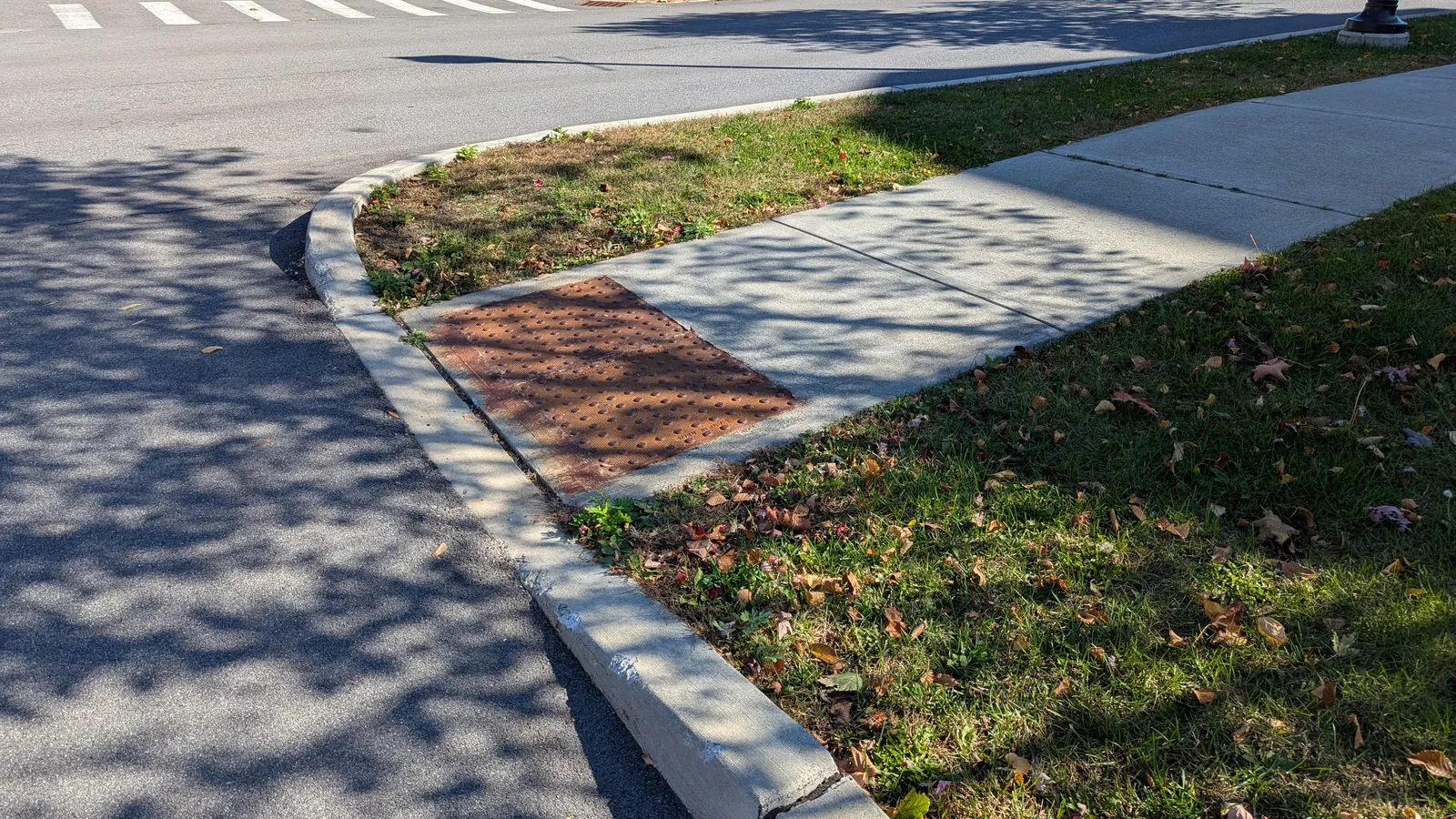 Concrete sidewalk ramp sloping down to a curb cut with a red-brown tactile warning panel at the street edge