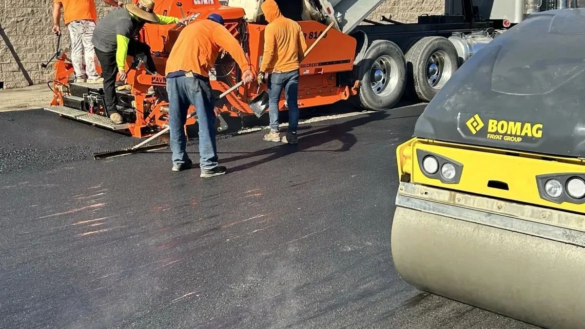 Paving crew raking fresh asphalt behind a paver with a steel drum roller in the foreground